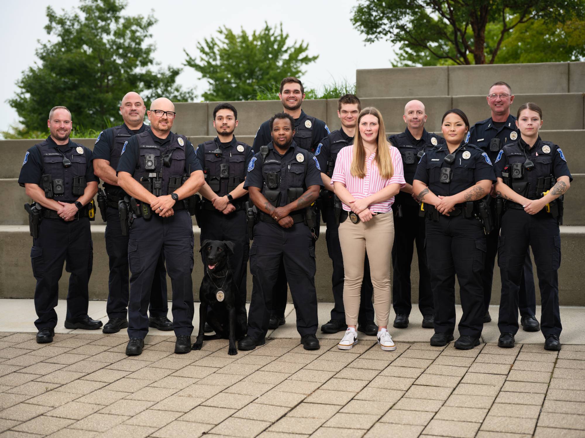 GVPD police officers in a staggered line, wearing their blue patrol uniforms, posing in front of the Student Services fountain. K9 dog Koda is laying in front. One officer, a detective is in plain clothes. The officers are smiling.